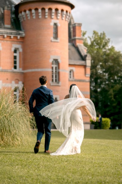 Bride has hand in the air walking hand in hand with groom on grass veil is blowing in the wind